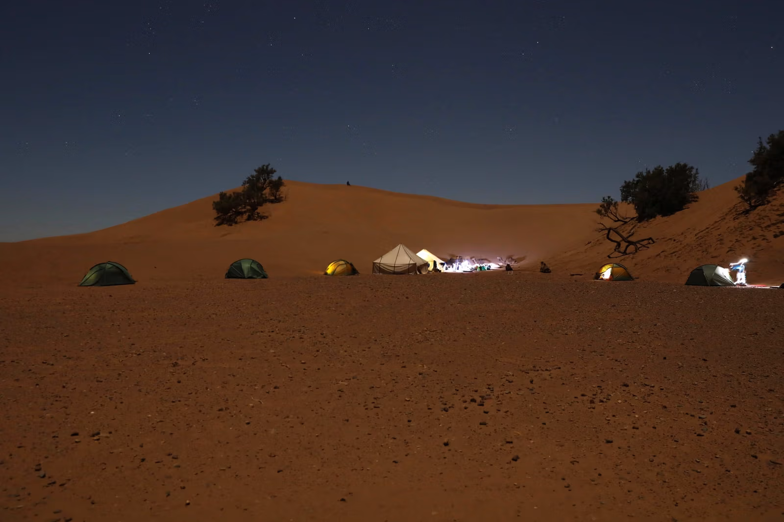 Camp lights glowing beneath a dune at dusk